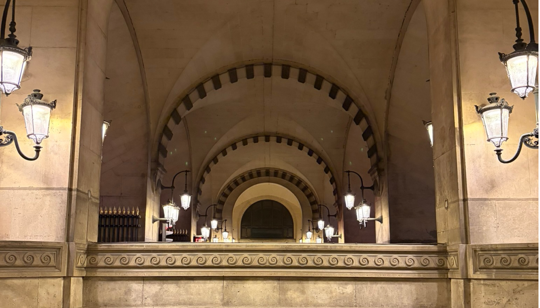 A corridor with arched ceilings and ornate stonework, lit by vintage-style lanterns on both sides.
