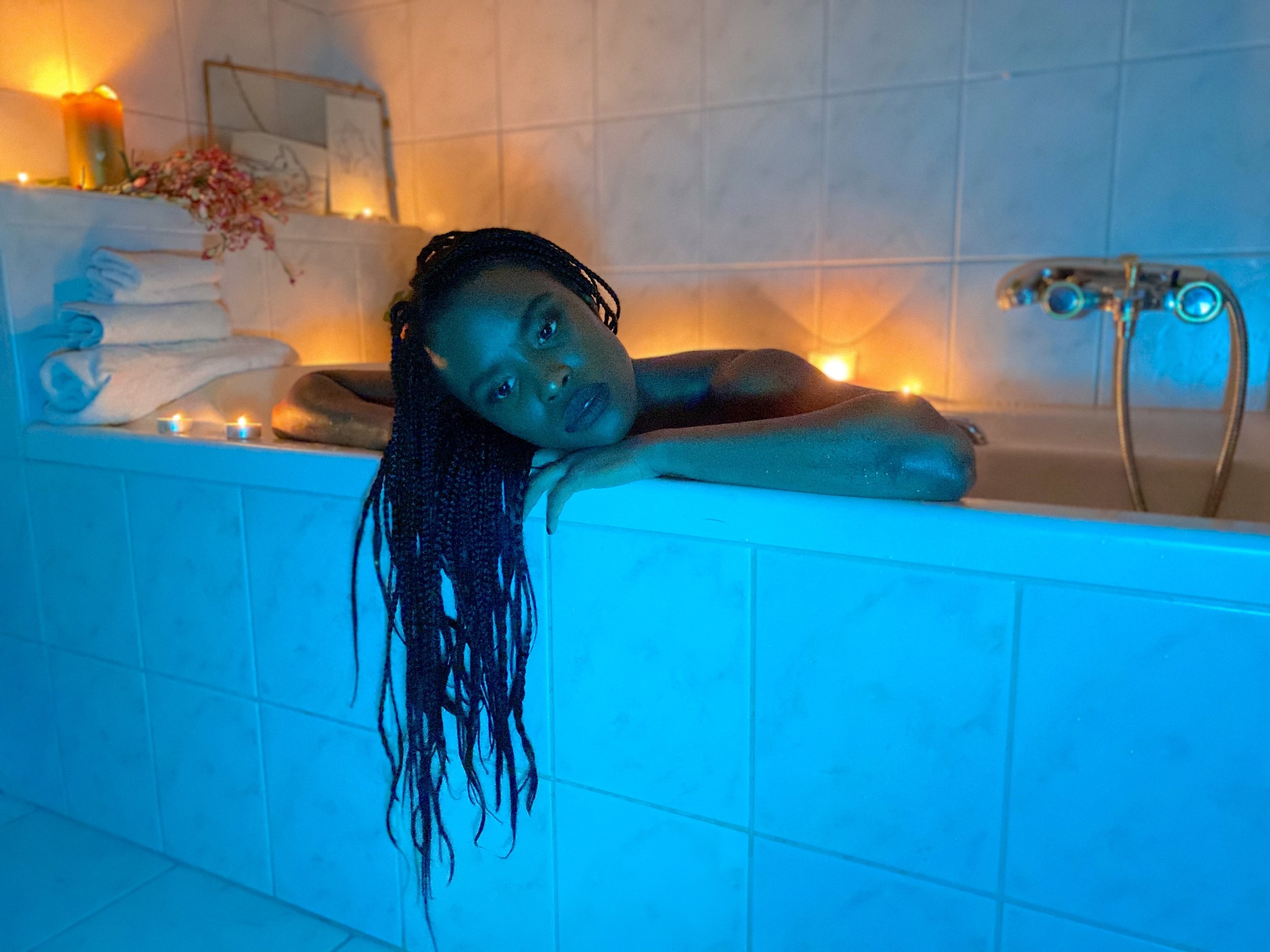 A woman with braided hair lying in a bathtub filled with water, looking at the camera. The bathroom has blue-tiled walls, and there are candles, a mirror, and a floral decoration on a shelf in the background, creating a cozy atmosphere.