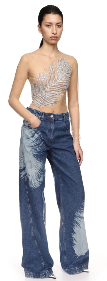 Fashion model wearing a sheer, long-sleeved top with a feather design and blue wide-leg jeans with feather print lasered on, posing with hands in pockets.