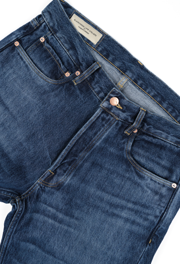 Close-up of dark blue denim jeans with orange stitching, showing the waistband, front pockets, and copper rivets.