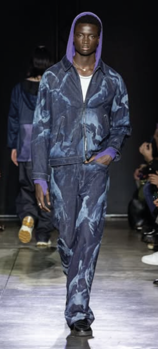 A male model walking on a fashion runway, wearing a matching denim laser patterned jacket and pants with a purple hoodie underneath, and sneakers.