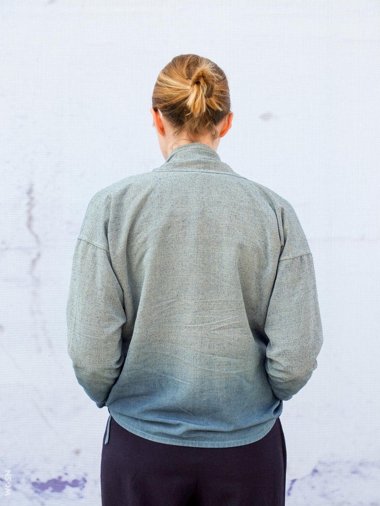 Back view of a person with blonde hair tied in a bun, wearing a gray zip-up jacket and dark pants, standing against a plain light background.