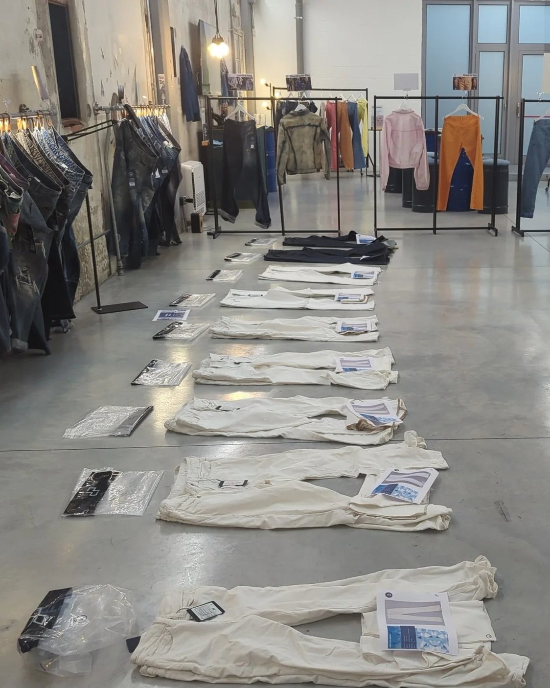 White pairs of pants laid out on the floor with informational sheets on top, in a clothing store or showroom with additional clothing on racks and a concrete wall in the background.