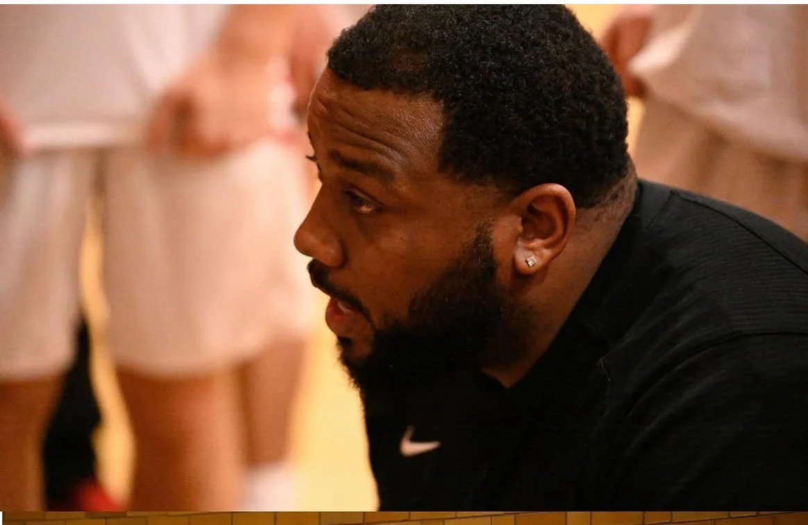 Close-up of a man with short curly hair, beard, earring, wearing a black Nike shirt, looking to the left.