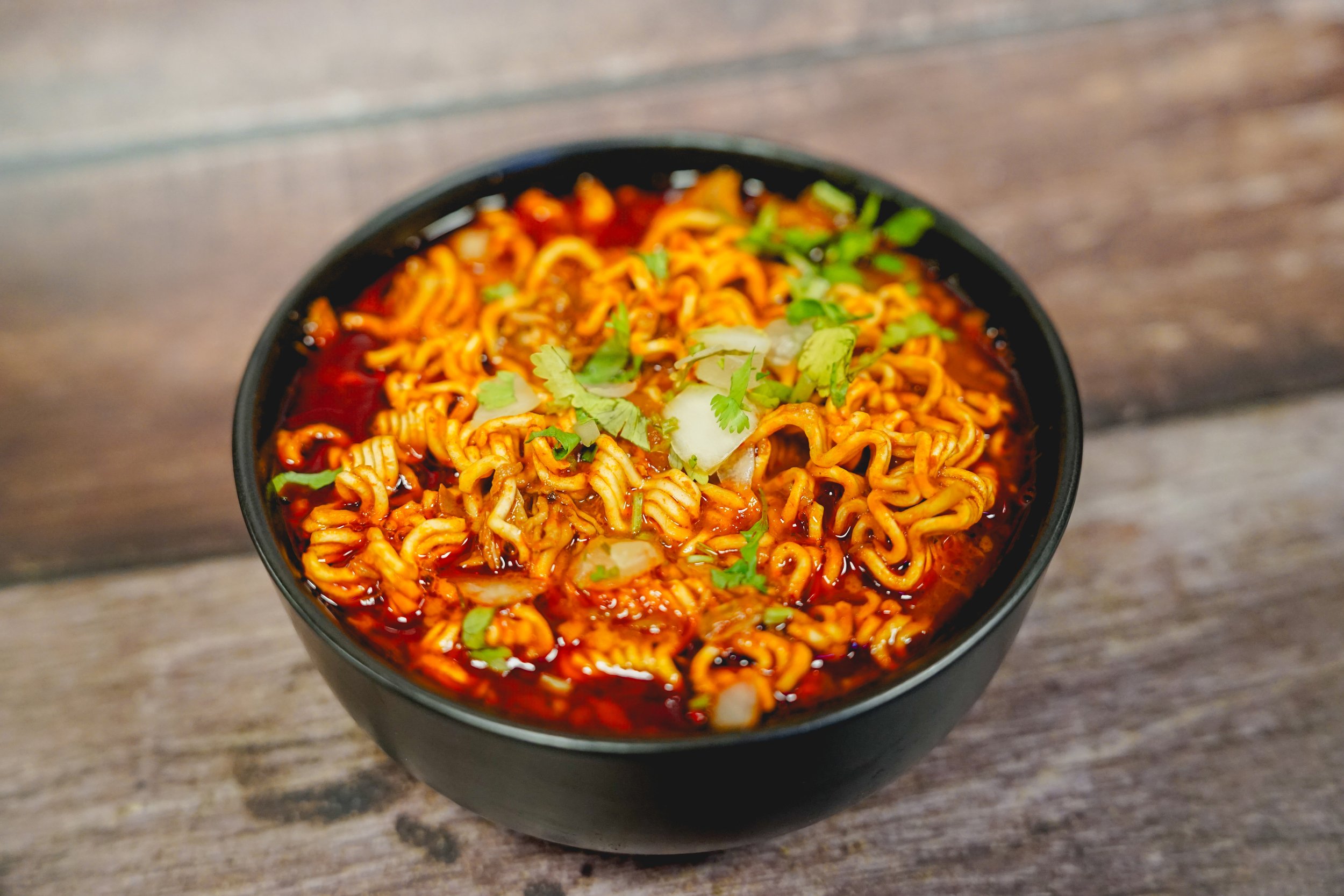 A bowl of spicy ramen noodles garnished with chopped green onions and cilantro on a wooden surface.