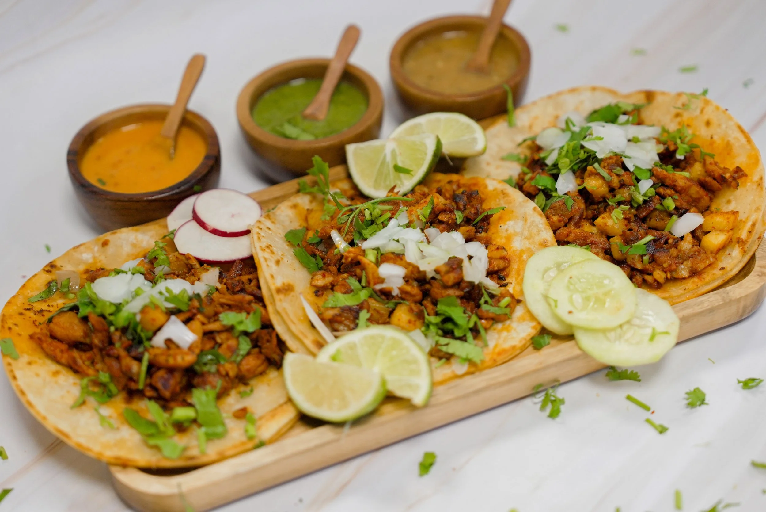 Close-up of three tacos with seasoned meat, chopped onions, cilantro, and sliced cucumbers on a wooden serving board, garnished with lemon wedges. Three small bowls of different sauces are in the background.