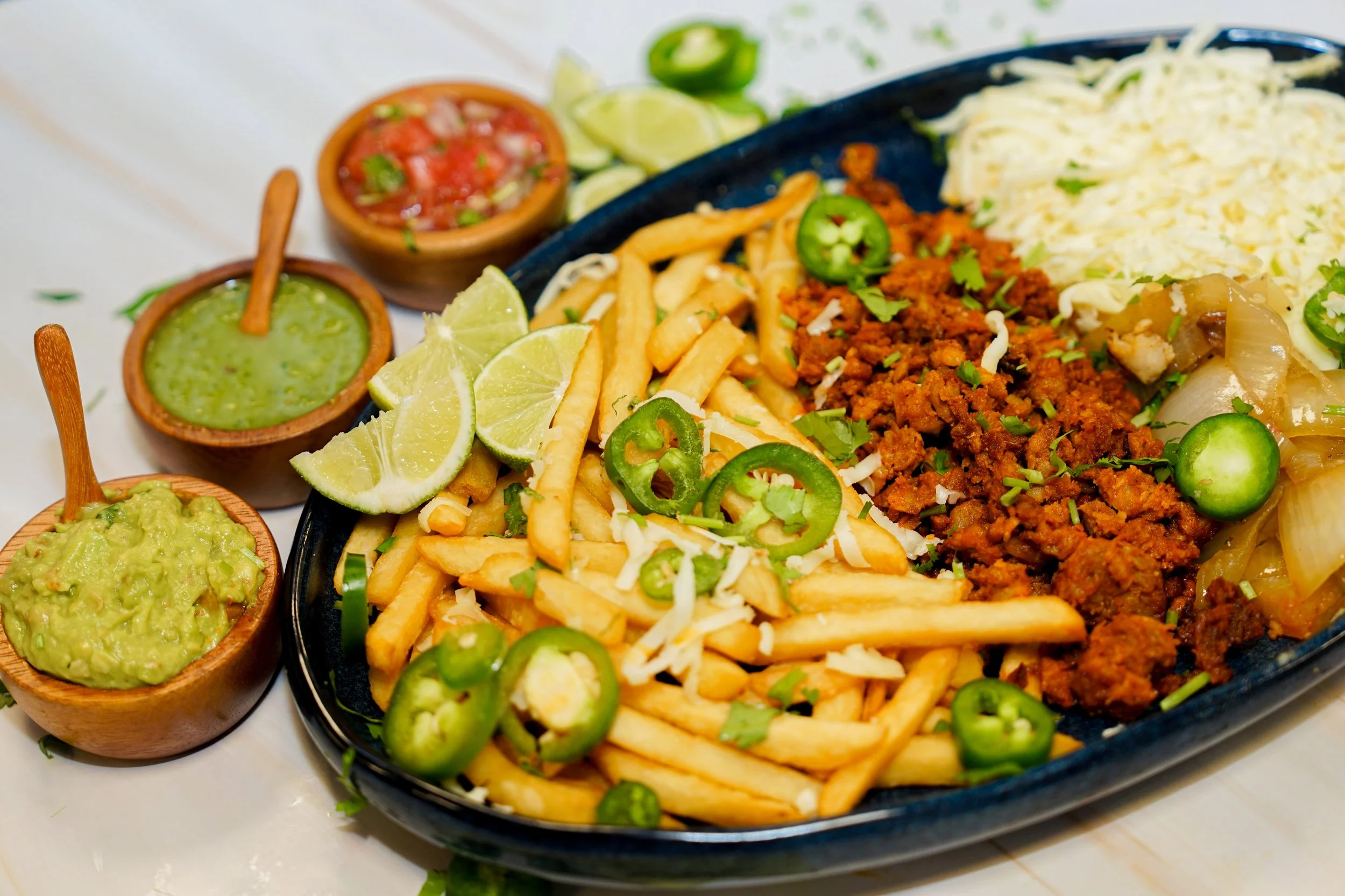 A black plate of Mexican food with seasoned ground meat, French fries, shredded cheese, jalapeño slices, and lime wedges, accompanied by small bowls of guacamole, green salsa, and red pico de gallo.