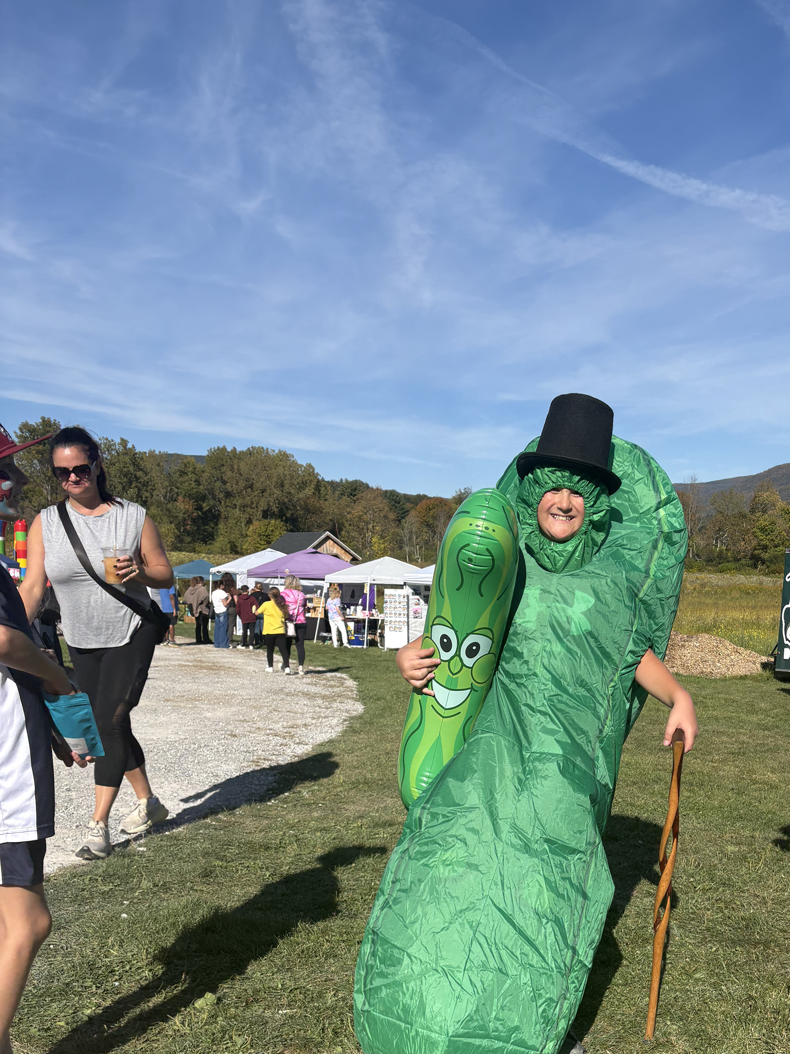 Person dressed as a green plant with a black top hat at an outdoor event, surrounded by tents and people, under a blue sky.