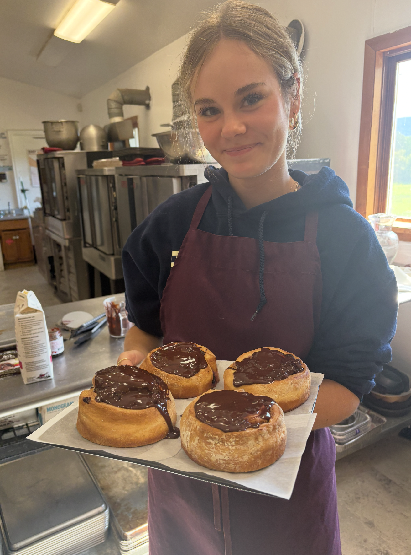 A young woman wearing a black hoodie and maroon apron holding a tray of four cinnamon rolls topped with chocolate glaze in a kitchen.