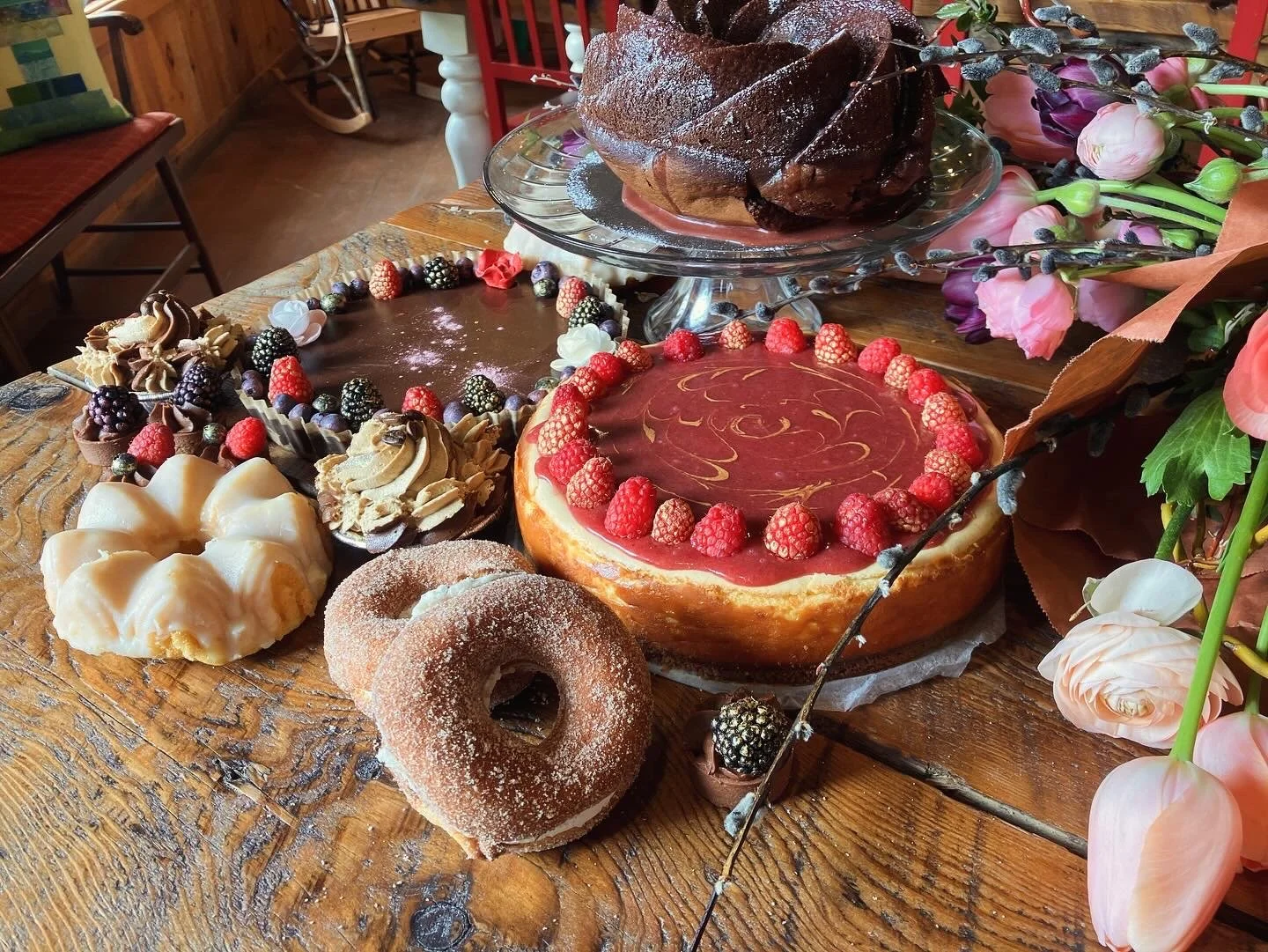 A wooden table displays various desserts including a donut with cinnamon sugar, a chocolate tart with berries, a cheesecake topped with raspberries, a layered mousse dessert, a fruit tart with raspberry and lemon swirls, and a chocolate cake on a glass cake stand. There are also decorative flowers surrounding the desserts.