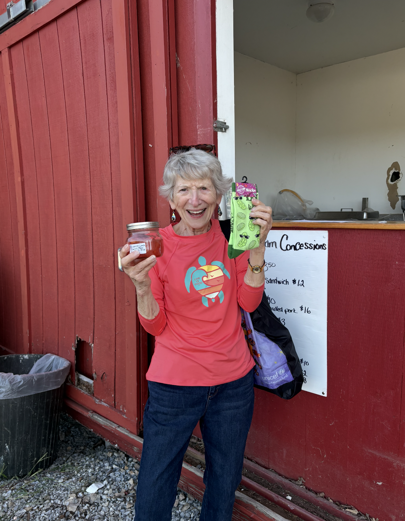 A woman with gray hair wearing a red shirt with a turtle logo stands in front of a red concession stand, holding a jar labeled 'Dianne Lamb Sweet Pickles' in one hand and a green sock in the other. She is smiling and wearing sunglasses on her head.