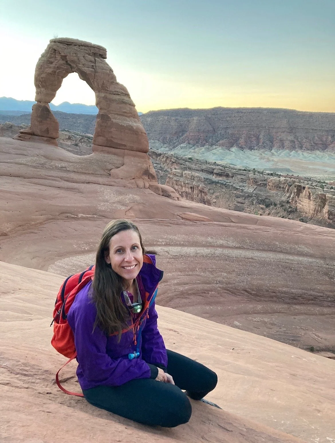 A smiling woman with long brown hair, wearing a purple jacket and black pants, sitting on desert rocks with a red backpack. Behind her is the Delicate Arch in Arches National Park, Utah, during sunset or sunrise.