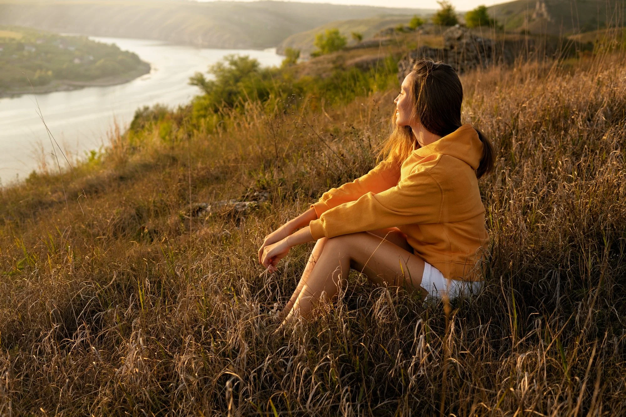 A woman sitting on a grassy hillside overlooking a river in a valley during sunset, wearing an orange hoodie and beige pants, with her eyes closed and enjoying the view.