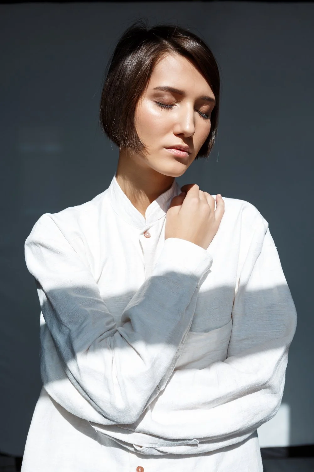 A woman with short brown hair, closed eyes, and wearing a white shirt, is standing with her hand on her shoulder, in front of a dark background.