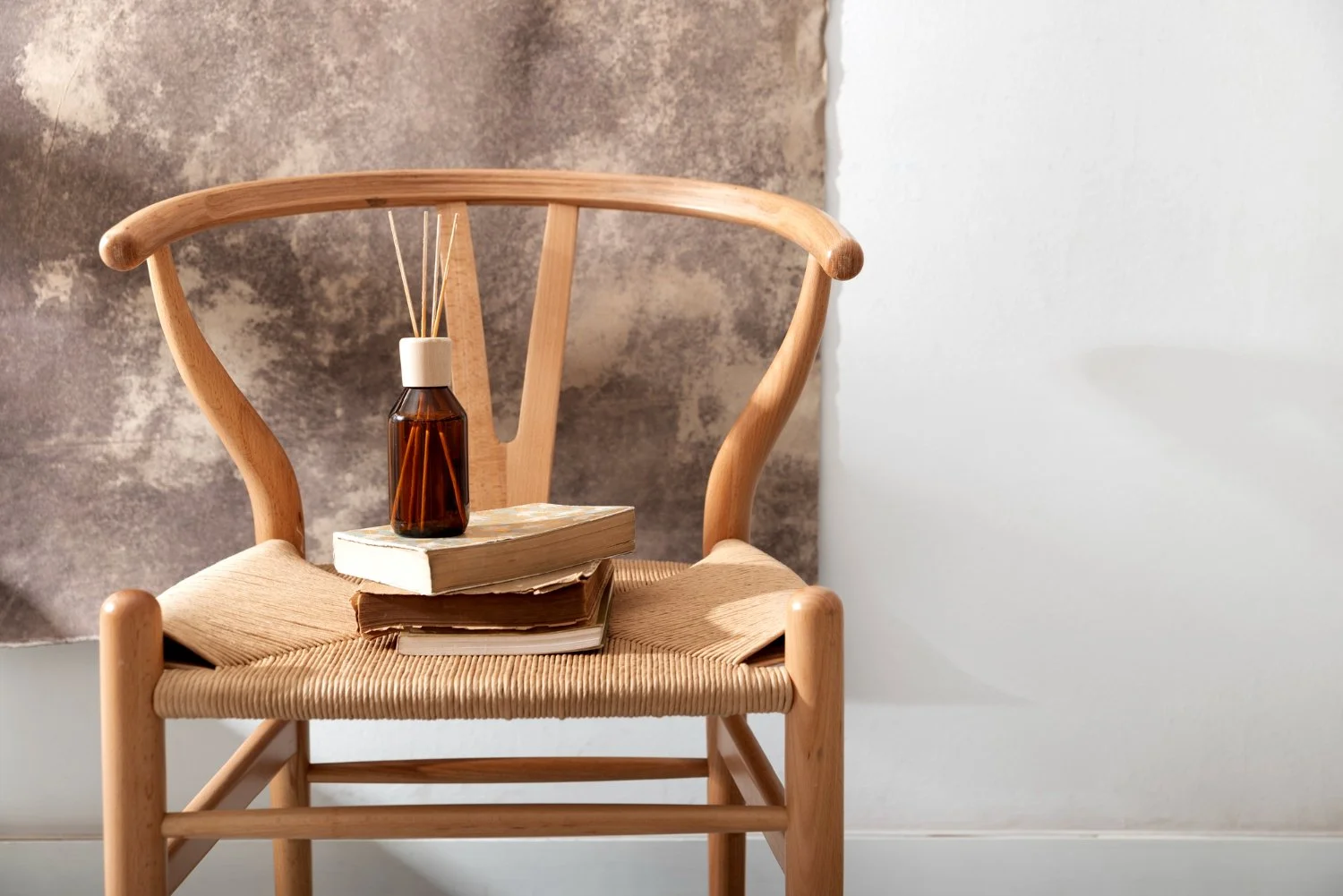 A wooden chair with a stack of three books on the seat, and a brown glass bottle with reed diffuser sticks on top of the books, set against a textured wall and plain white wall.