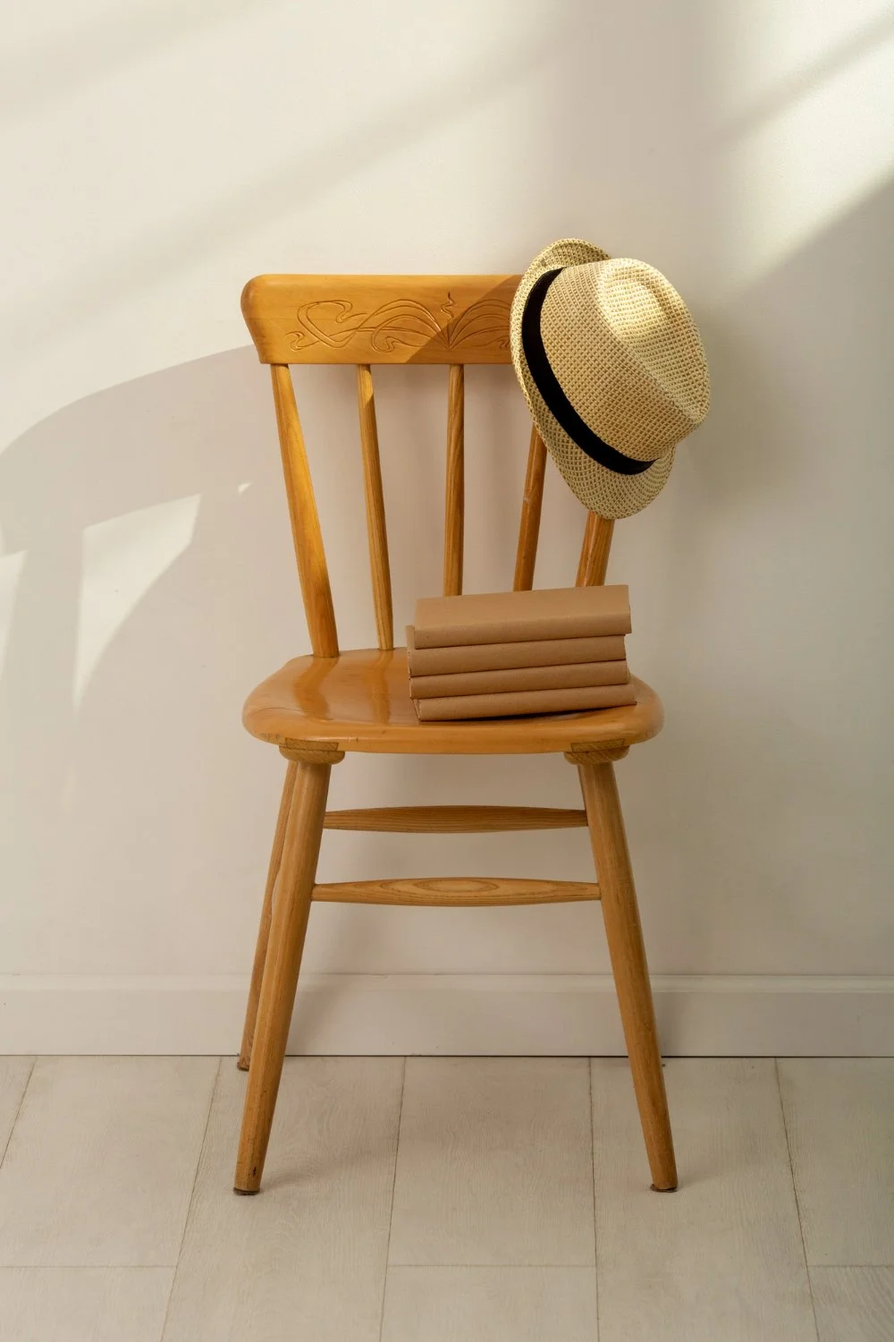 A wooden chair with a carved backrest against a white wall. A straw hat with a black band hangs on the chair's backrest. Four brown notebooks stacked neatly rest on the seat.