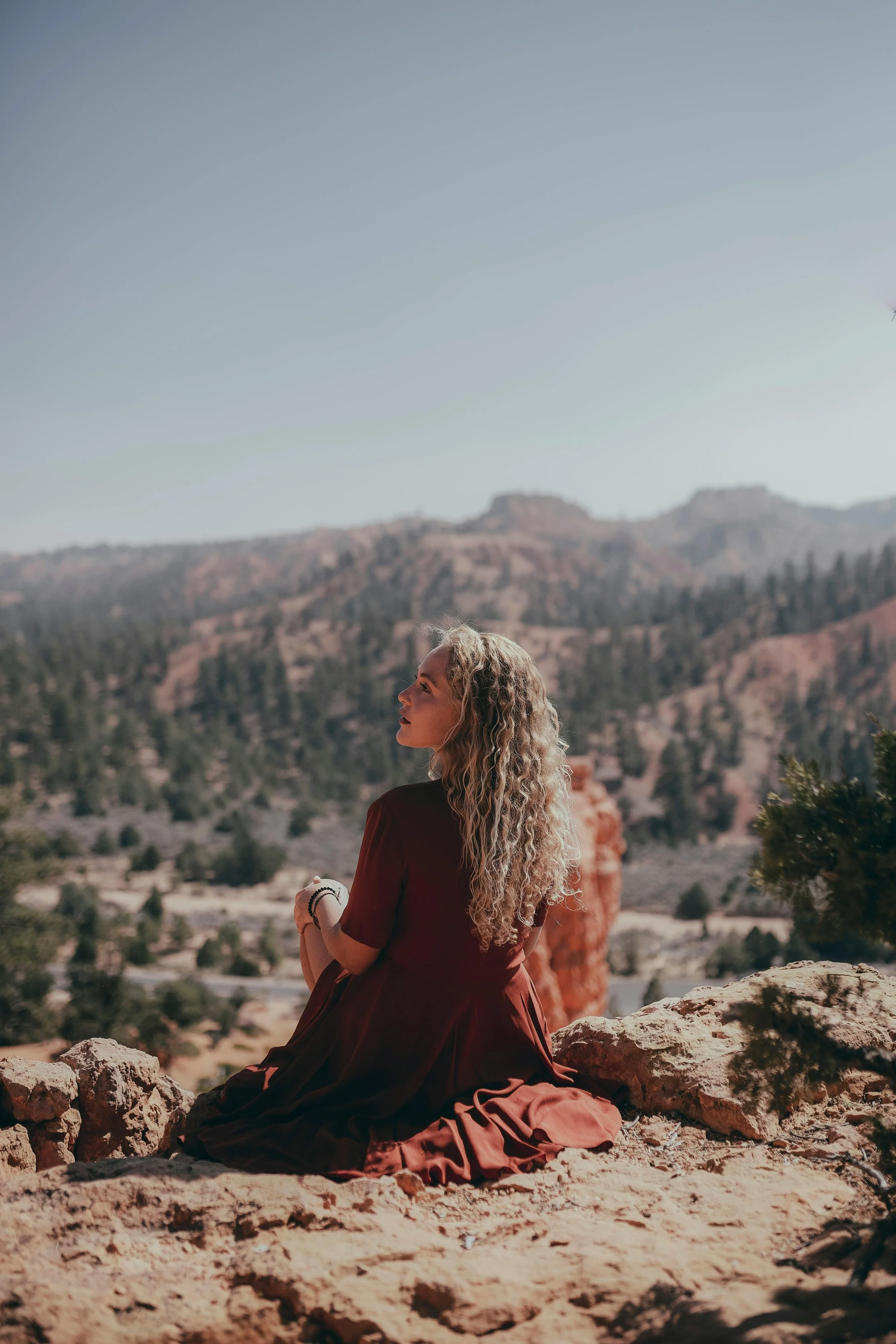 A woman with long curly hair sitting on rocky terrain with a mountainous landscape and clear sky in the background.
