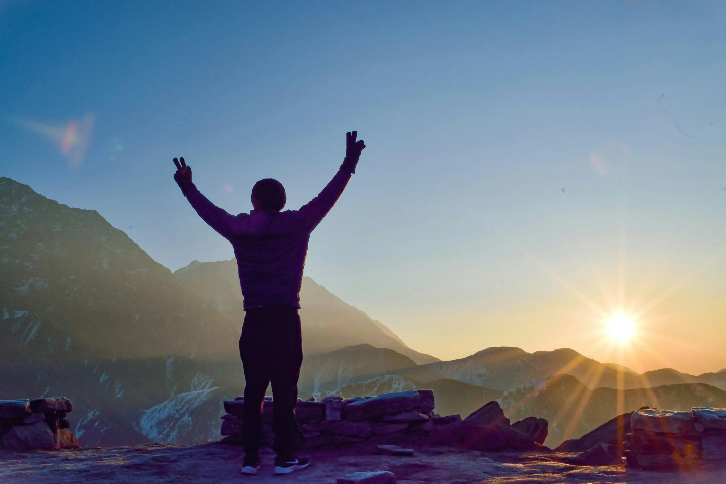 Person standing on rocks with arms raised, making peace signs, at sunset in a mountainous landscape.