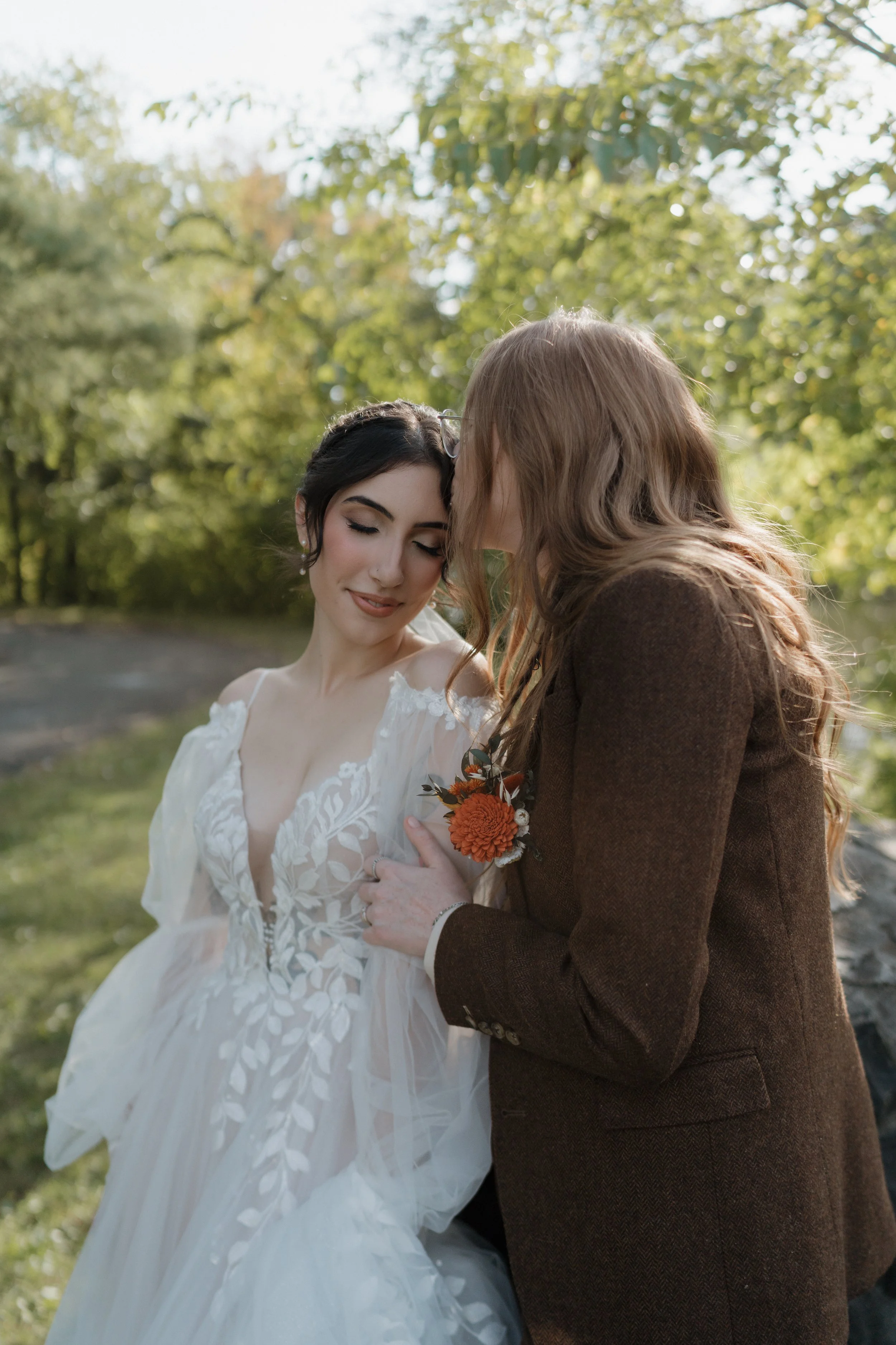 A bride with dark hair and a white dress and a woman with long red hair wearing a brown blazer share an intimate moment outdoors surrounded by green trees.