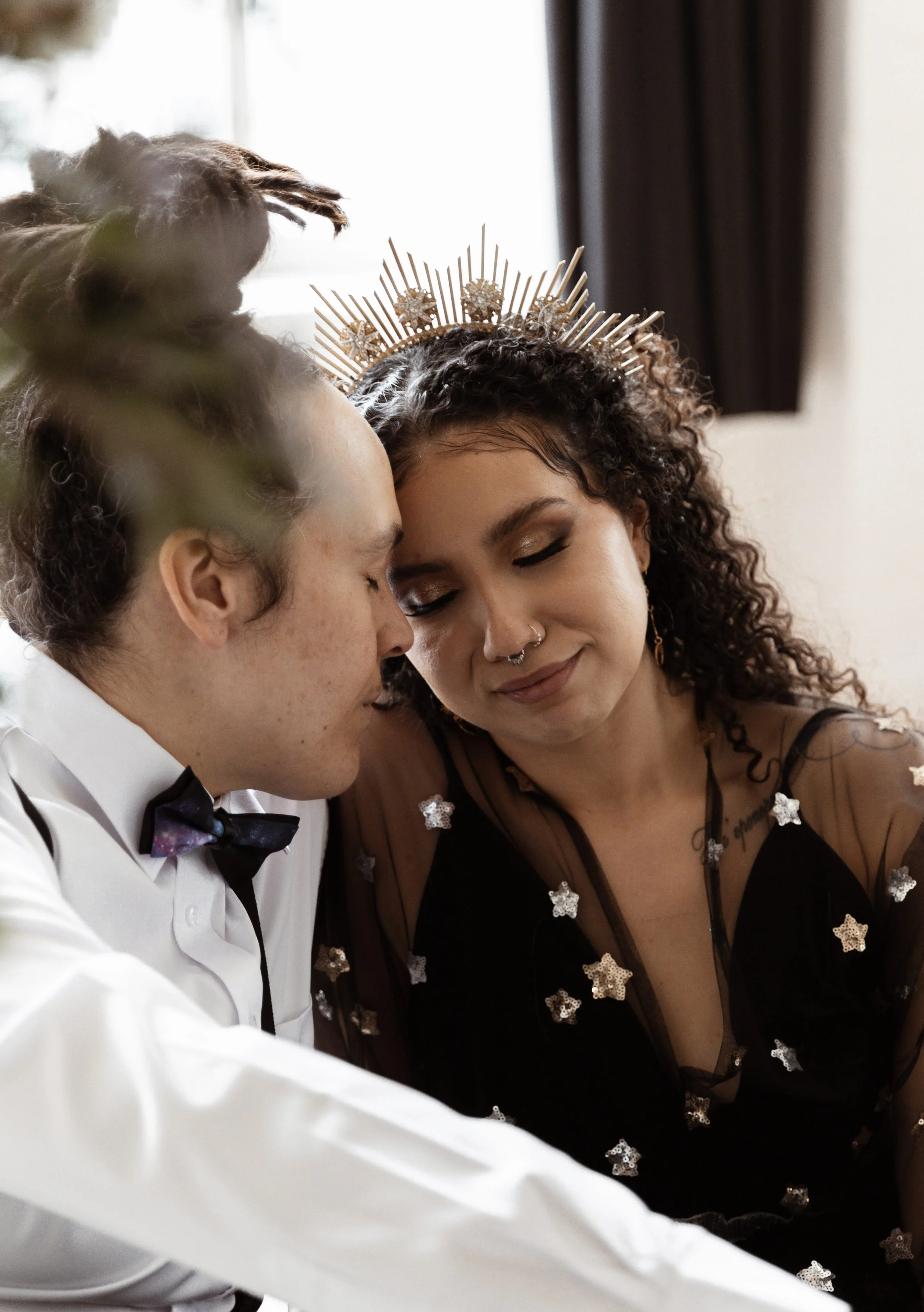 Two women with their foreheads touching, one wearing a crown and the other in a tuxedo, sharing a tender moment indoors.