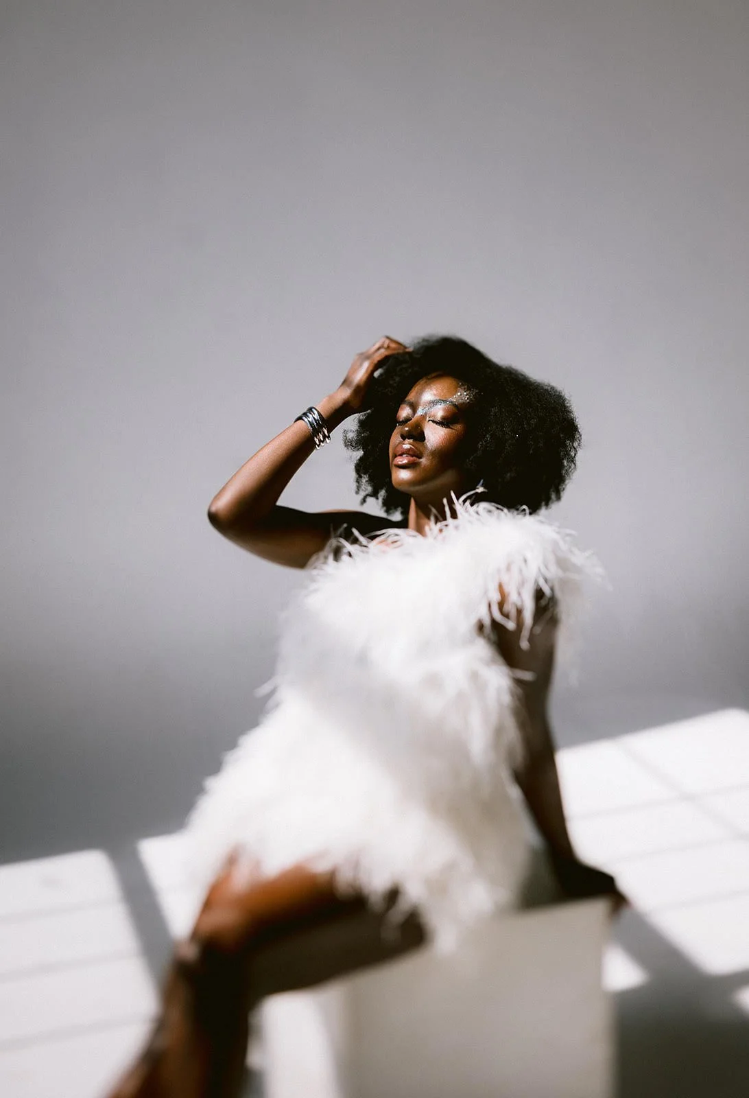 A woman with dark curly hair wearing a white feathered dress poses with eyes closed and one hand on her head against a plain gray background.