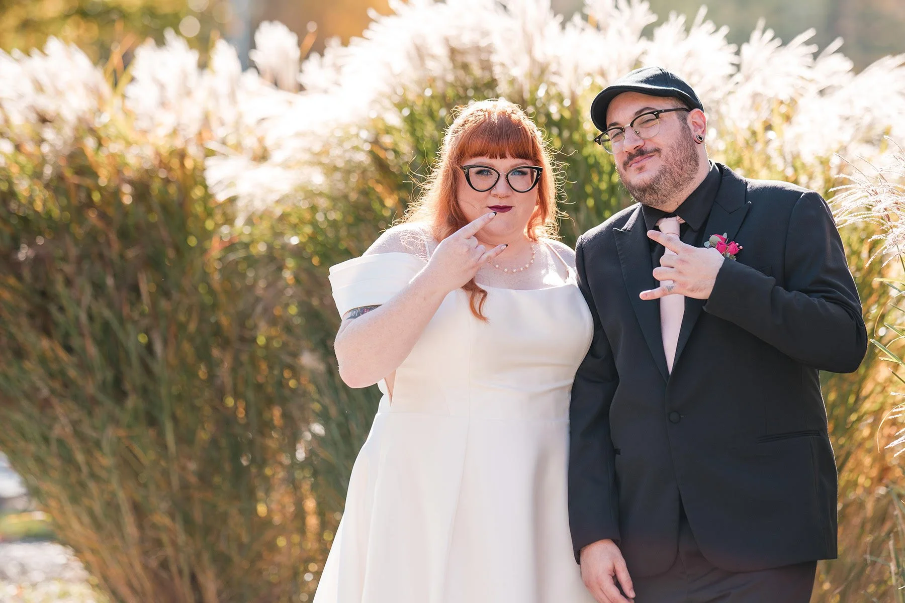 A couple standing outdoors at a wedding, both making rock and roll hand signs and smiling.