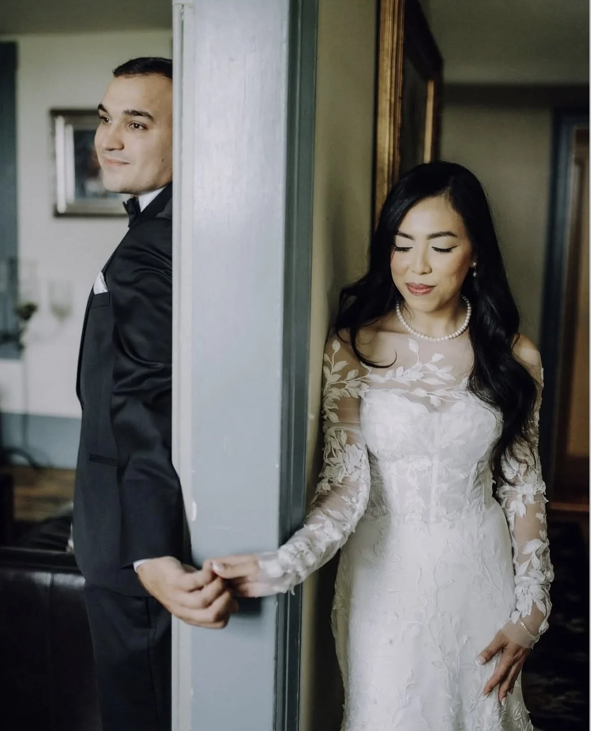 A groom in a black tuxedo stands on one side of a wall, looking away smiling, while a bride in a white lace wedding dress with pearl necklace and earrings stands on the other side, looking down with a gentle expression, holding hands behind the wall.