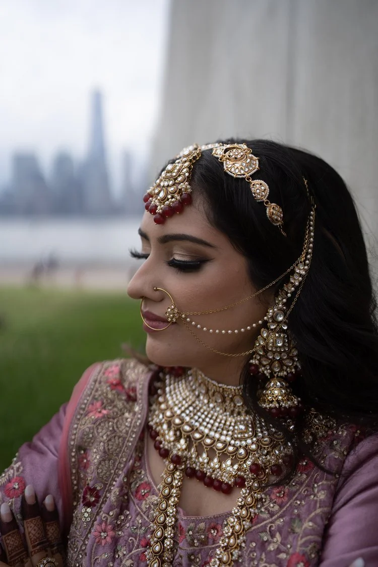 A woman in traditional Indian bridal attire with elaborate jewelry, including necklaces, earrings, a nose ring, and a headpiece, standing outdoors with a cityscape in the background.