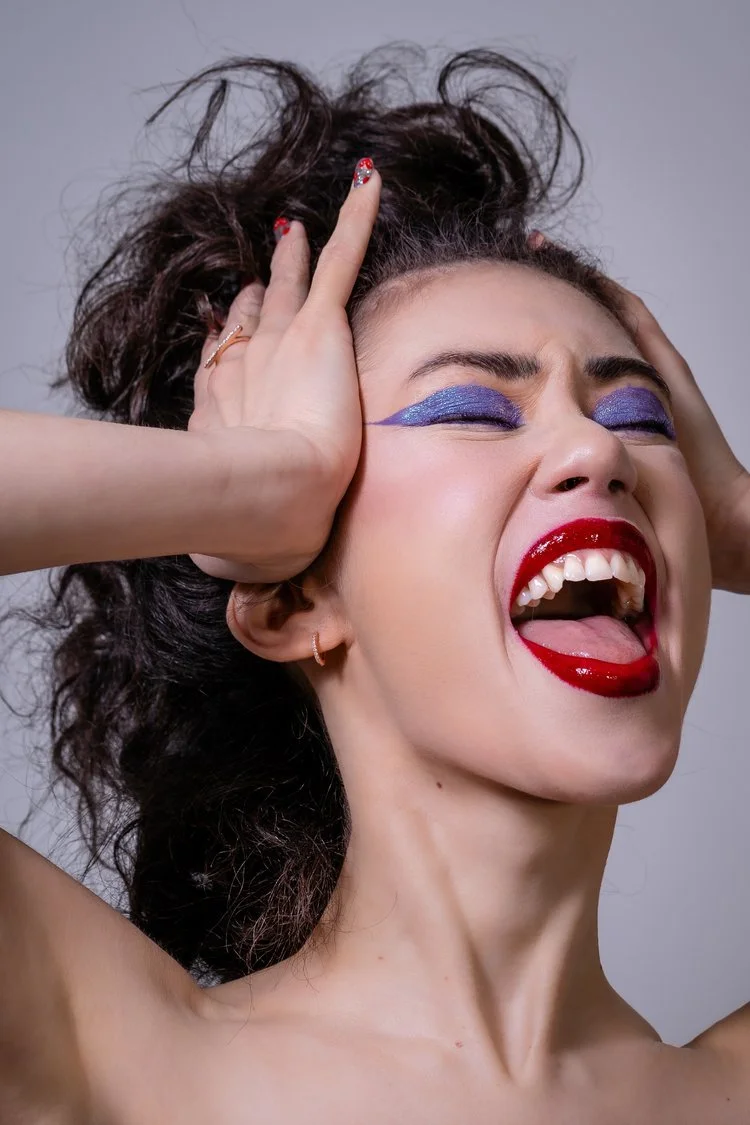 A woman with wild, curly hair and bold makeup, including purple eyeshadow and bright red lipstick, holding her head with both hands and expressing frustration or anguish.