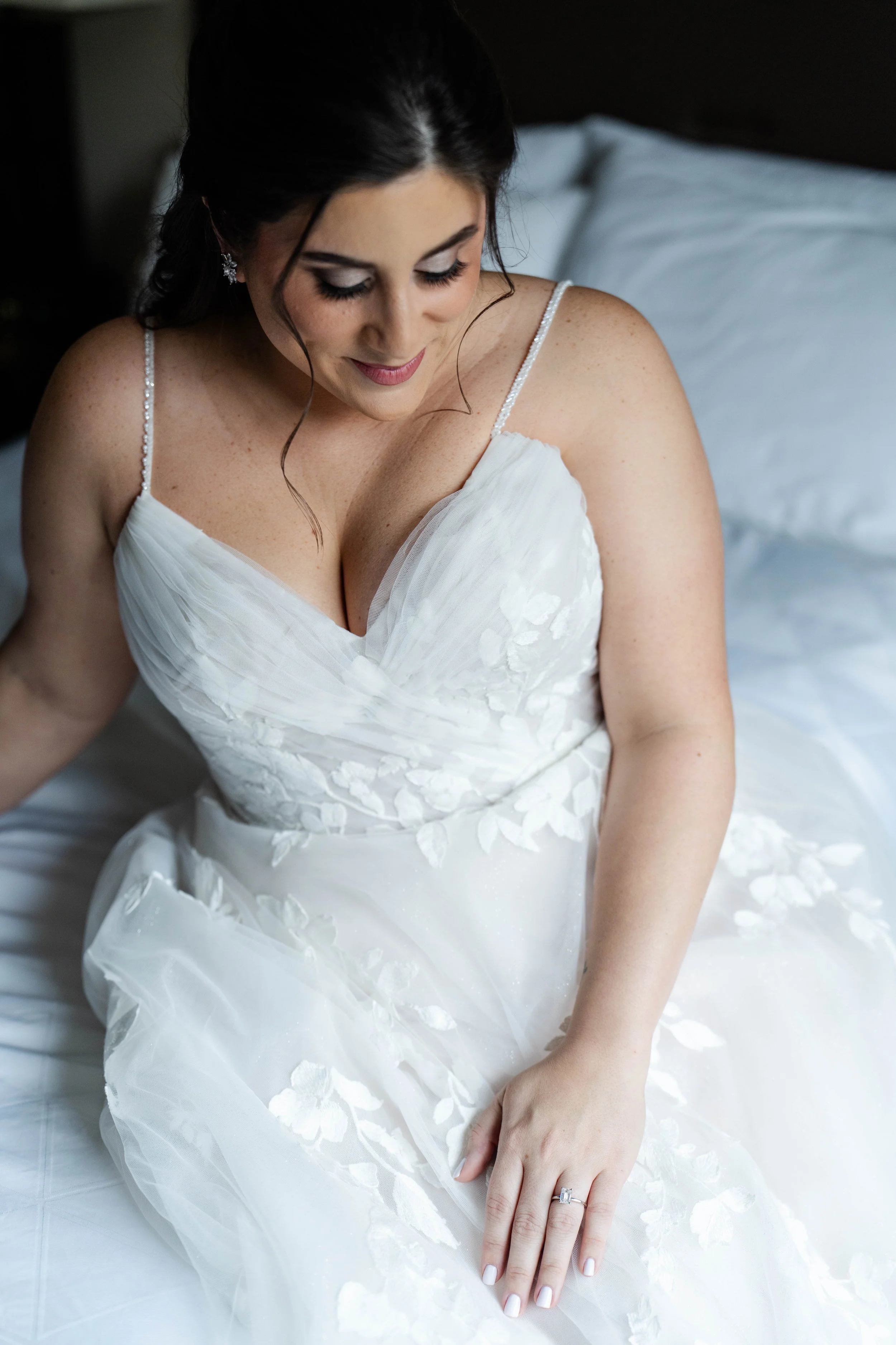 A bride in a white wedding dress with spaghetti straps, sitting on a bed, looking down with a gentle smile, with dark hair styled in loose waves, wedding ring visible on her left hand.