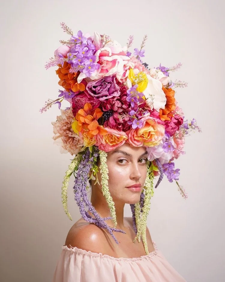 A woman wearing a large headdress made of various colorful flowers, including purple, pink, orange, and white blooms, with some hanging down like tassels, in front of a plain light background.
