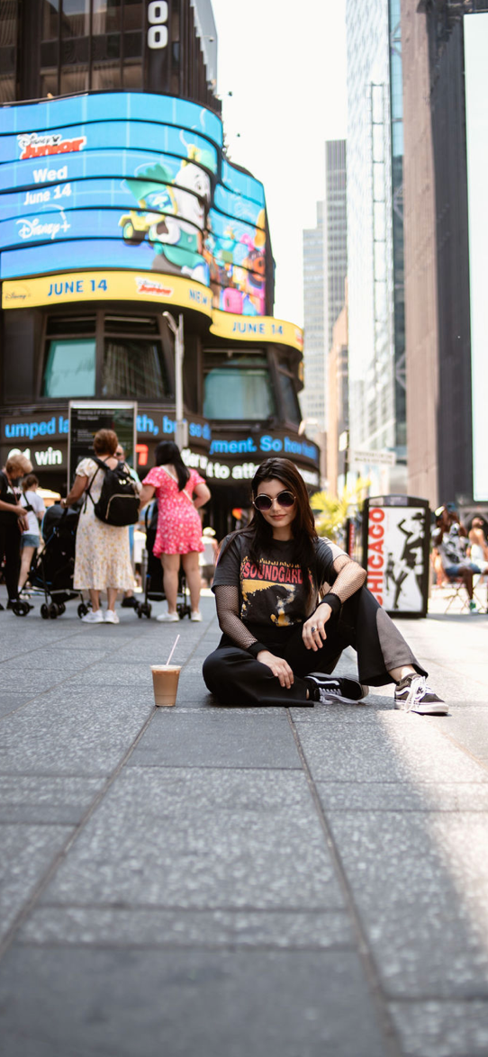 A young woman sitting on the sidewalk in Times Square, New York City, with a beverage on the ground beside her. She is wearing sunglasses, a dark t-shirt, and black pants, with tall buildings and a digital billboard in the background.