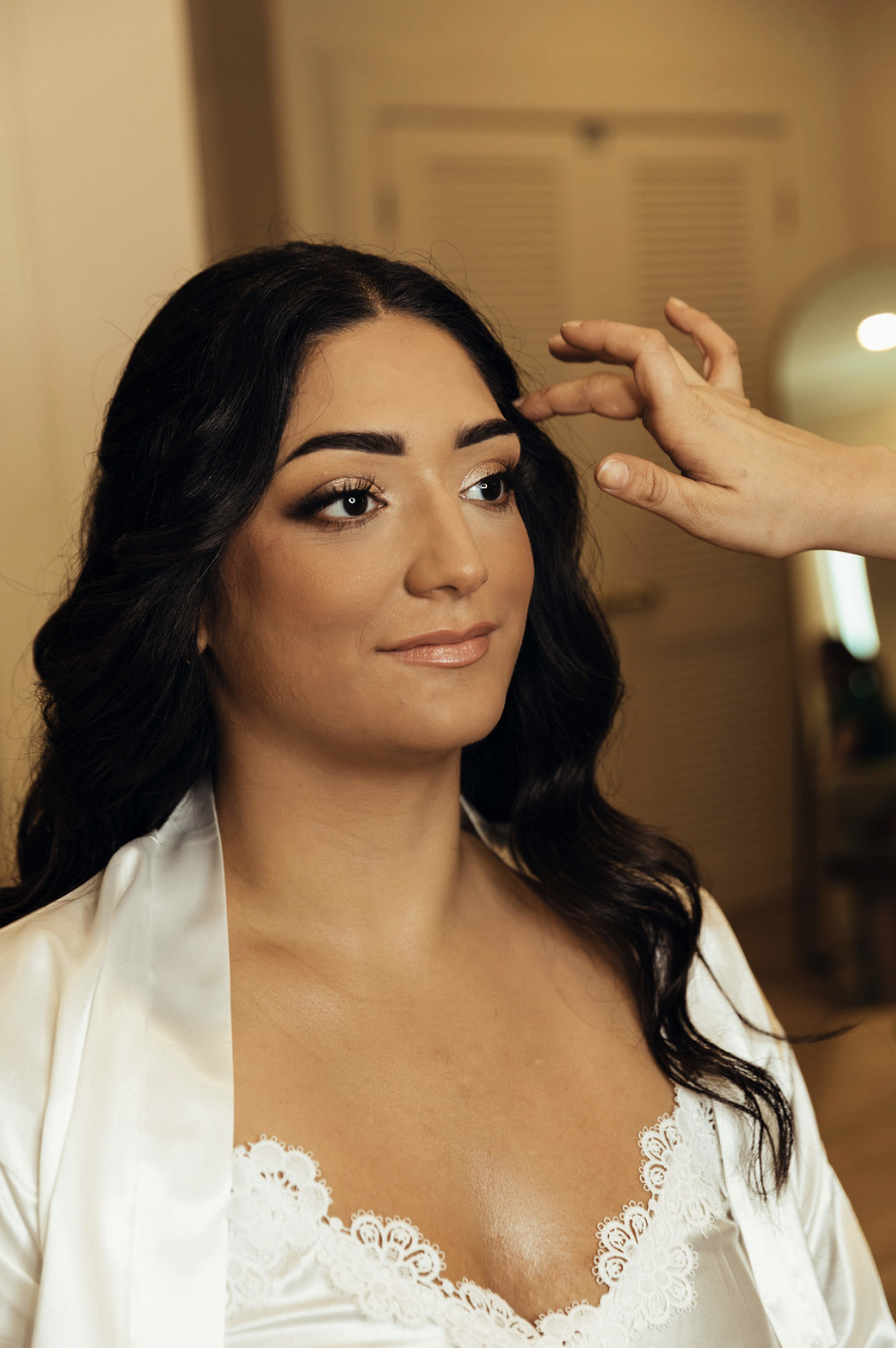 A woman with dark, wavy hair and makeup sitting as a makeup artist applies makeup or adjusts her hair, wearing a satin robe with lace trim. The background is warm with soft lighting.