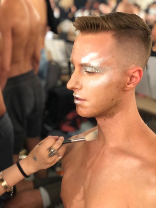 A shirtless man getting makeup applied to his face by a makeup artist.