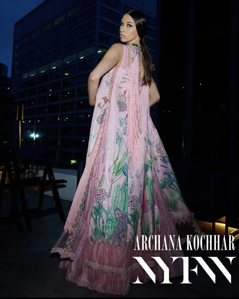 Woman in a pink embroidered dress with floral pattern, standing in a cityscape at night, with text 'Archana Kochar NYFW' on the image.