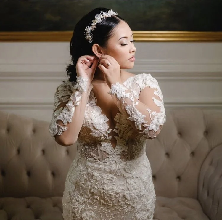 Brunette bride in a lace wedding dress adjusting her earring in a room with beige furniture.
