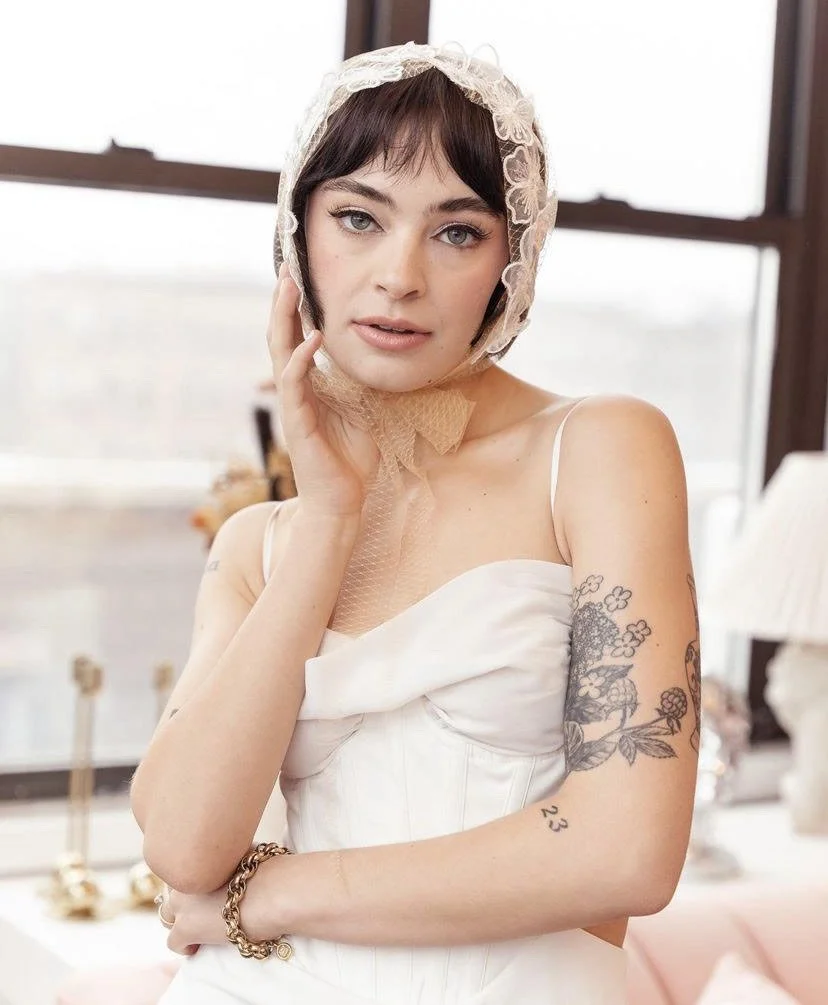 A young woman with short dark hair and tattoos on her right arm, wearing a white dress and a lace bonnet, posing indoors with a city view in the background.