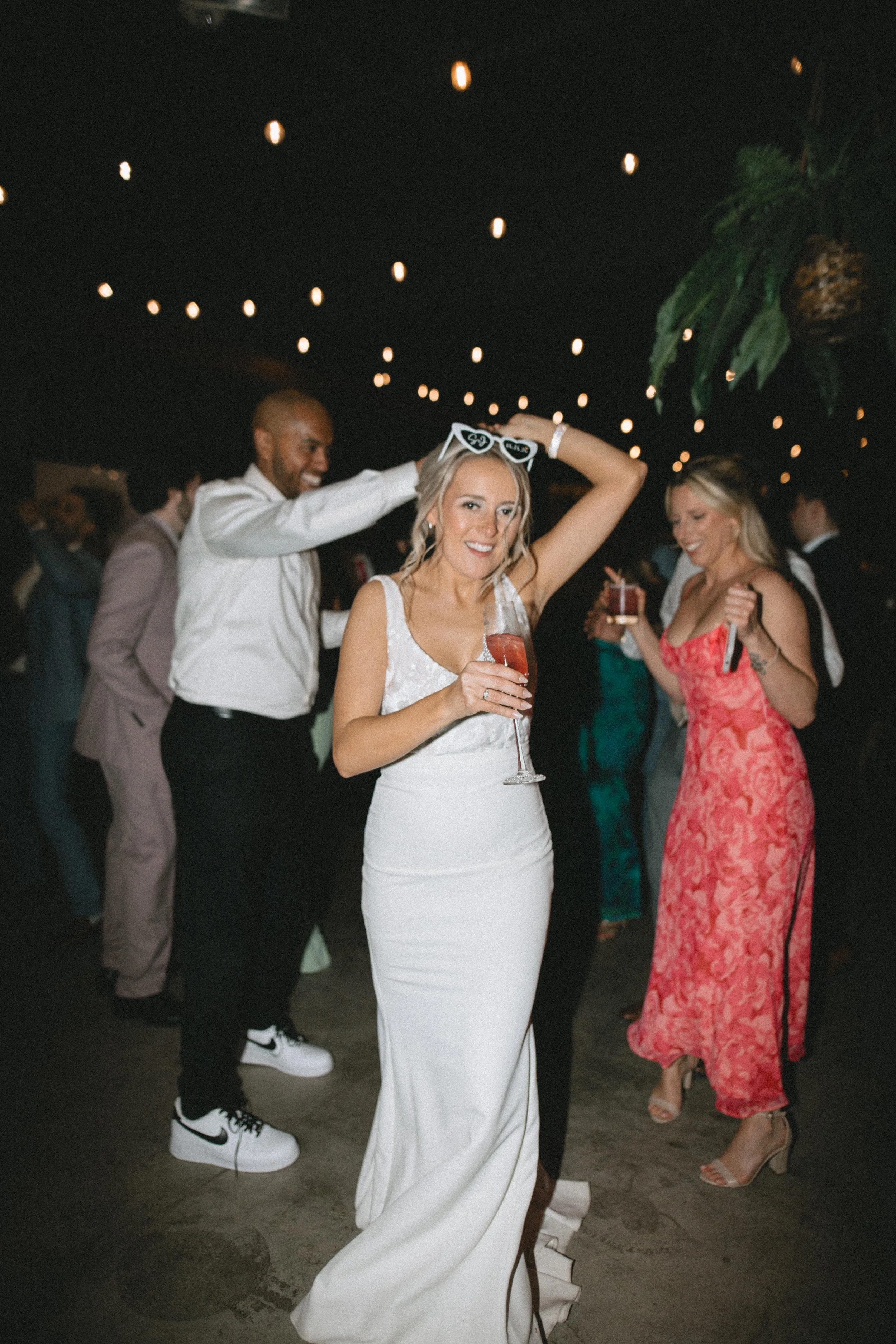 A group of people dancing at an event. A woman in a white dress is holding a drink and smiling. A man behind her is adjusting her glasses with a big smile. Other women and men are dancing in the background, some holding drinks. The ceiling is decorat