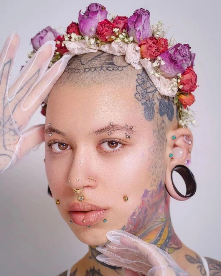 Close-up of a woman with numerous facial piercings, tattoos, and a floral headpiece made of pink, purple, and red roses. She is wearing transparent gloves and has varied piercings in her ears and face.