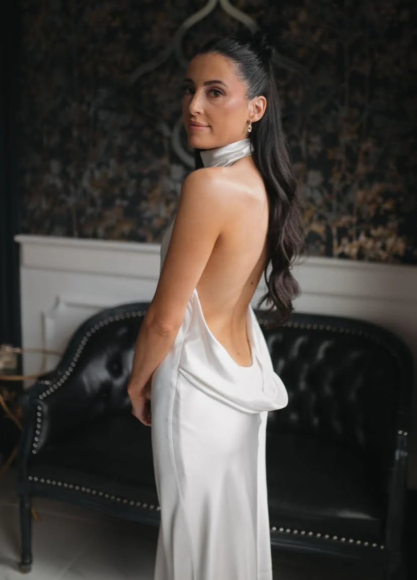 A woman in a satin wedding dress with an open back, standing in an indoor setting with a dark patterned wall and black leather couch behind her.