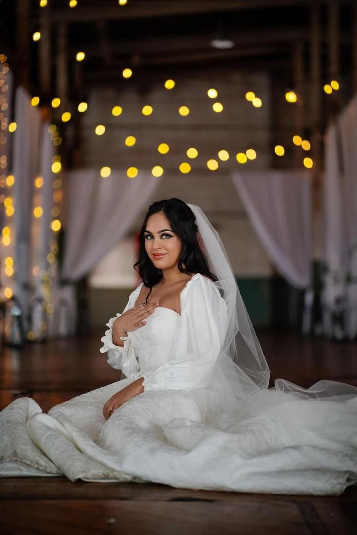 A bride in a white wedding dress sits on the floor inside a decorated venue with string lights and white drapery.