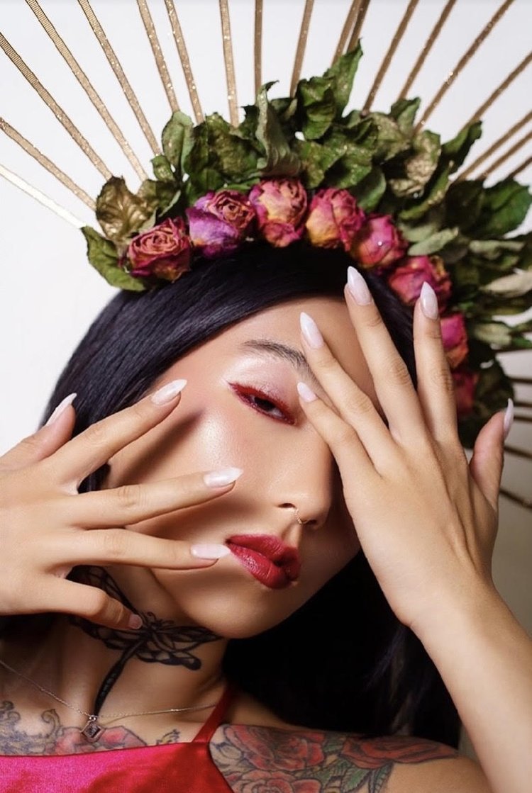 A woman with long black hair wearing makeup with red lipstick and eyeliner, a floral crown of dried roses, and a red top, making a fashion pose with her hands.