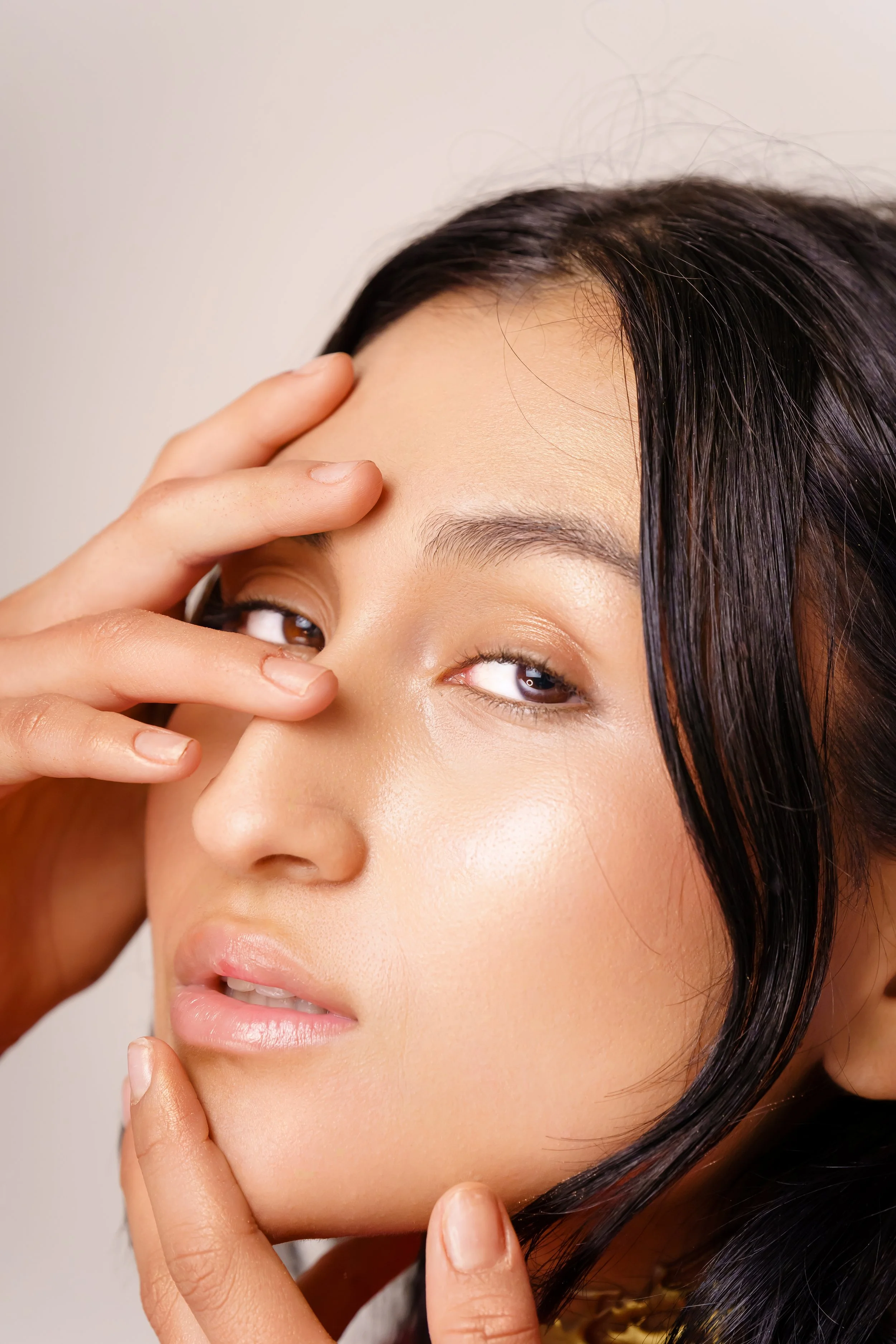 Close-up of a woman with dark hair, holding her face with her fingers, partially covering her eyes, with smooth skin and natural makeup.