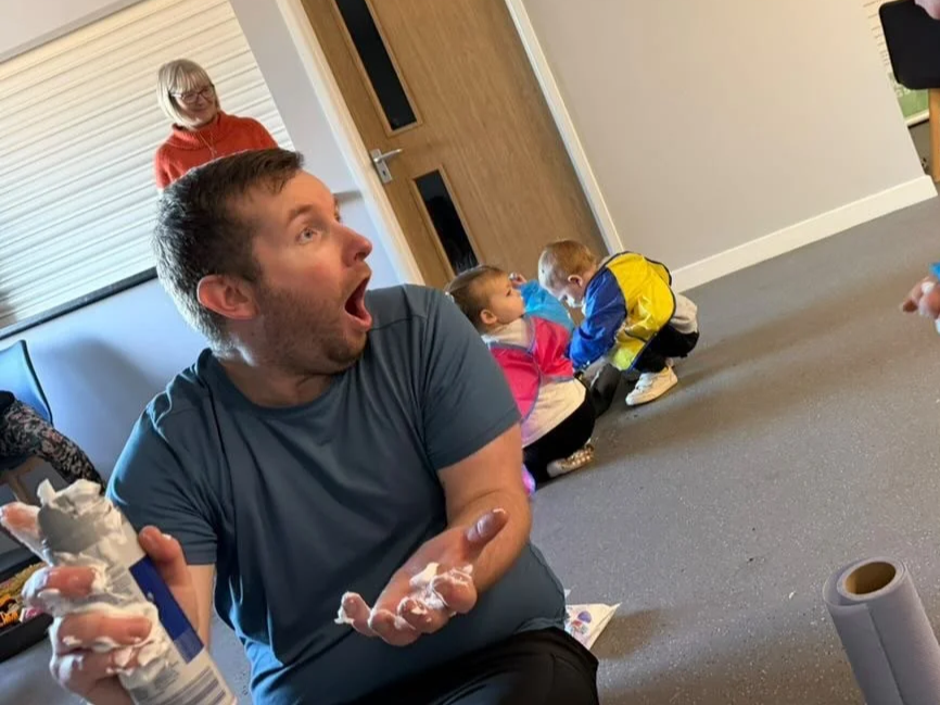 Man with shaving cream on hands and shirt reacting with surprise, in a room with children playing on the floor and a woman standing in the background.