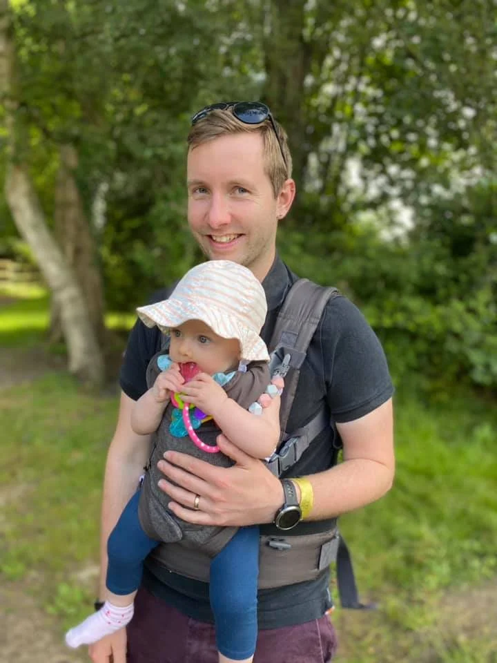 A smiling man wearing sunglasses and a smartwatch is holding a baby in a baby carrier in a park. The baby is wearing a sun hat, blue leggings, and is chewing on a toy while looking to the side. Trees and greenery are in the background.