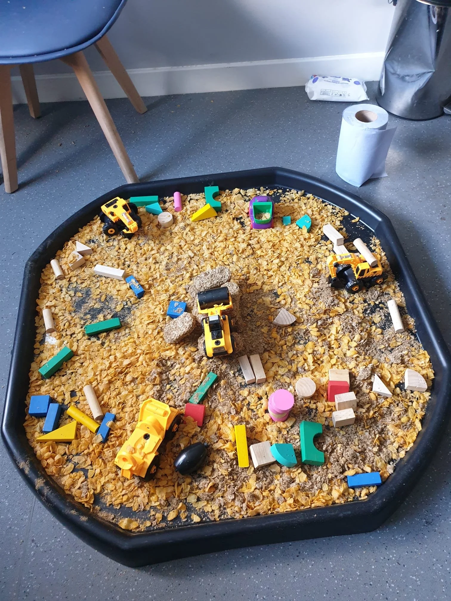 A play area with a black sandbox filled with yellow cornflakes and various toy construction vehicles, blocks, and figures. In the background, there is a blue chair, a roll of paper towels, a tissue pack, and a shiny metallic trash can.