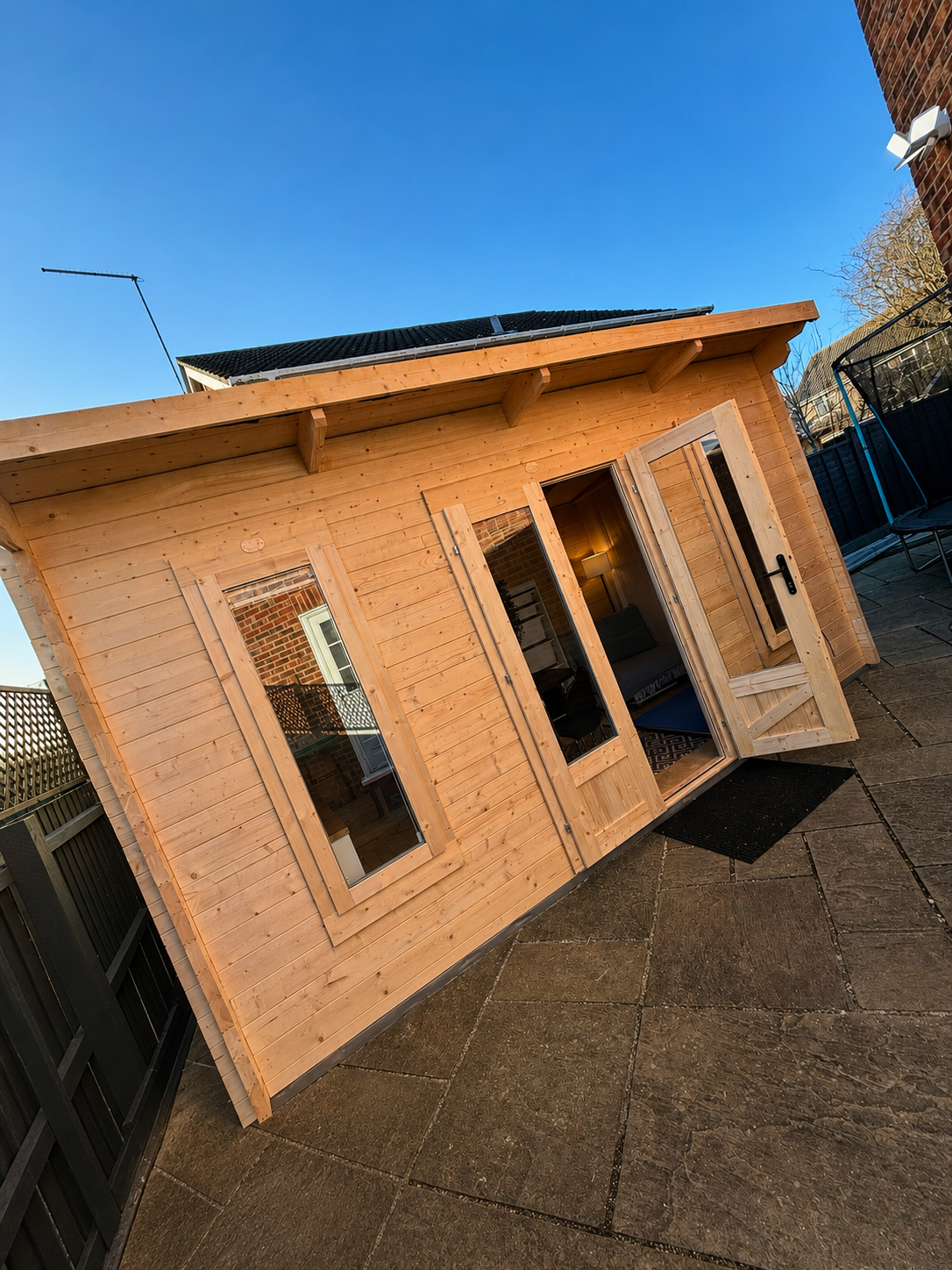 A small wooden garden house with windows and an open door, situated on a paved patio with a clear blue sky above.