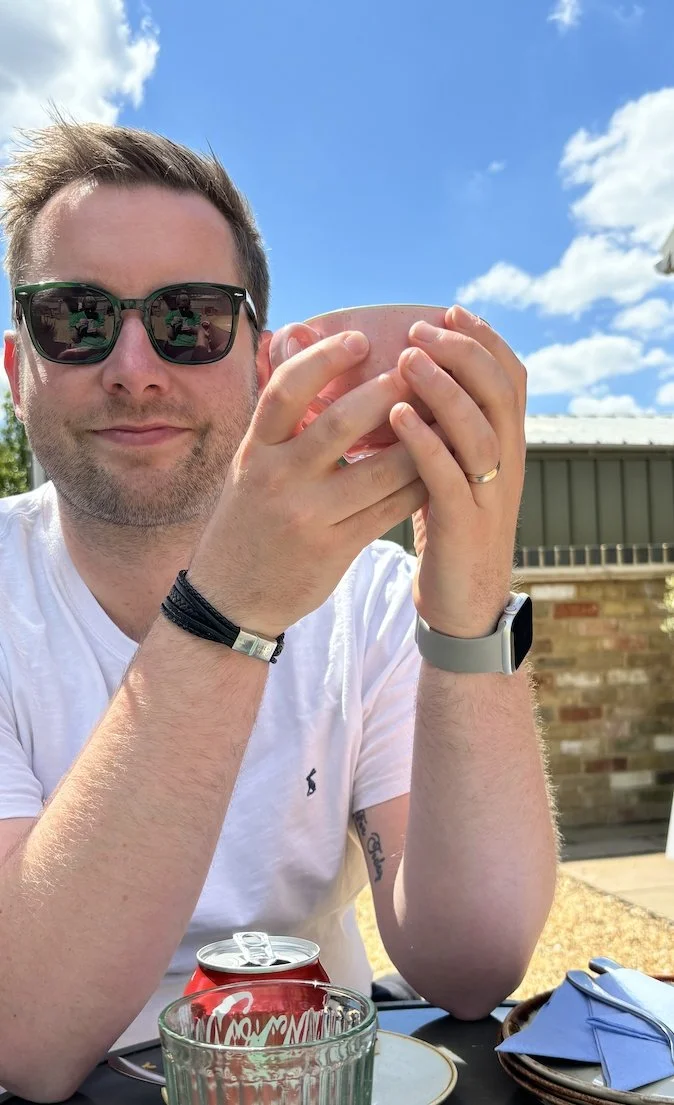 Man outdoors, wearing sunglasses and a white t-shirt, holding a pink mug, with a Coca Cola can and a glass on the table, blue sky with clouds in background.