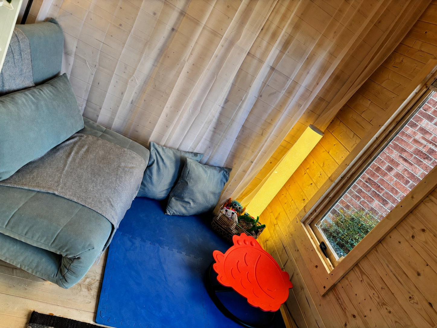 A cozy corner in a wooden room with a green couch, blue foam mat, red fish-shaped children's table, a basket of toys, a tall yellow lamp, and a window with a brick wall visible outside.