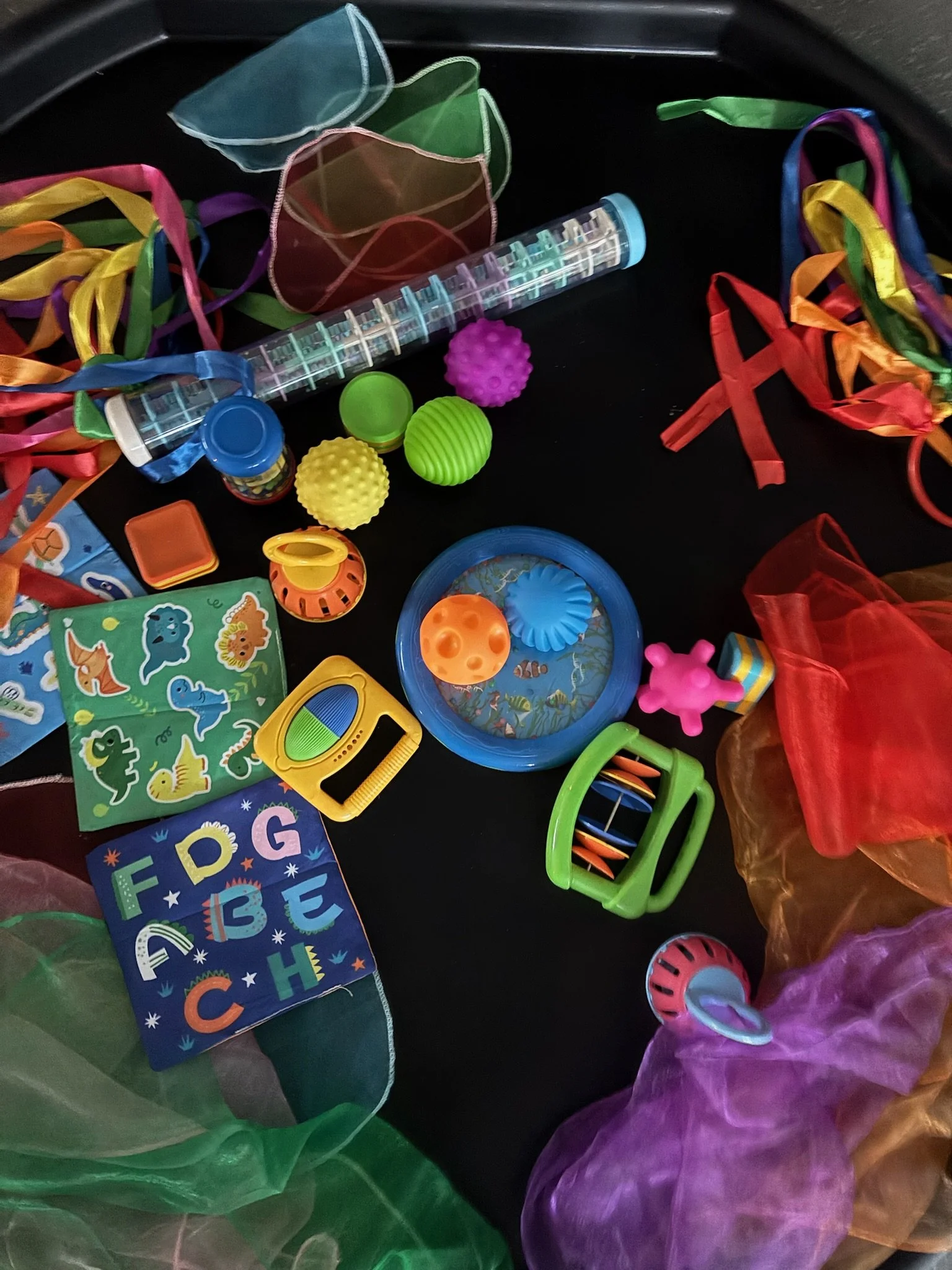 Colorful children's toys and educational materials on a black surface, including a plastic container filled with small multicolored beads, rubber balls, a toy drill, a colorful shape sorter, and a flap book with animal pictures.